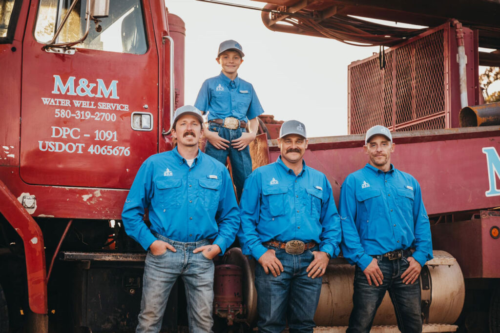 Three adult workers and a young boy from the M&M Water Well Service team pose in front of their red drilling truck. All dressed in vibrant blue uniforms and caps, they display a mix of pride and camaraderie. The boy, standing on the truck, adds a touch of familial warmth to the group portrait at dusk
