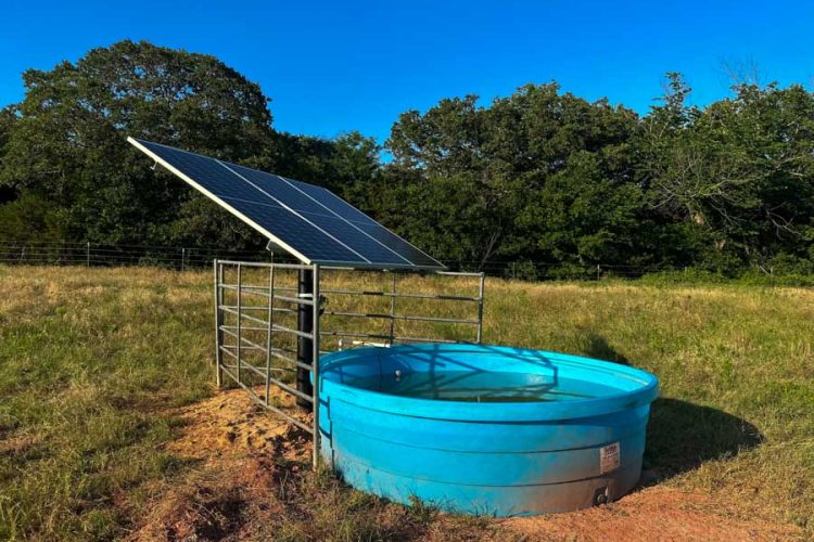 Solar-powered livestock watering system with blue tank in a rural pasture, installed by M&M Water Well Service