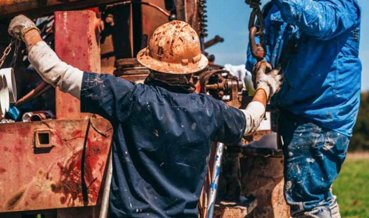 Two workers collaborate to secure a PVC pipe onto a drilling rig. One stands on the rig platform, handling the hook and chain, while the other assists from the ground. The scene, featuring various pipes and machinery, showcases the teamwork and precision required in the well drilling process, set against a clear sky in a rural environment.