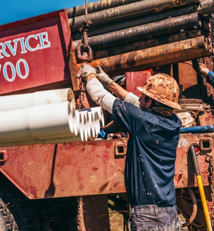 A worker in a hard hat and gloves secures a PVC pipe with a hook and chain to the drilling rig. The background shows other drilling equipment and pipes, highlighting the meticulous work involved in the well drilling process. The clear blue sky and rural setting emphasize the outdoor nature of the job.
