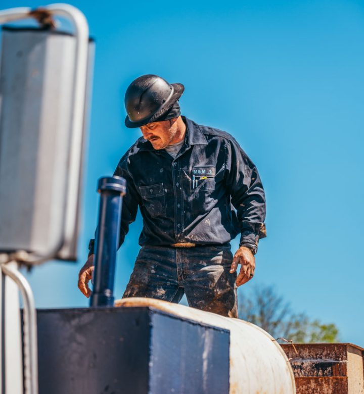 A focused water well driller, dressed in a black uniform and hard hat, stands on a drilling truck. He is managing a large cylindrical piece of equipment, possibly preparing for a drilling operation. The clear blue sky in the background enhances the outdoor work environment.