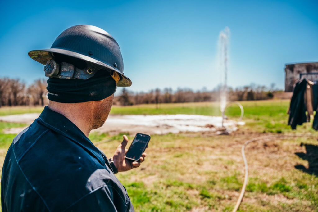 A worker in protective gear, including a hard hat, stands in a grassy field, holding a phone and monitoring drilling operations. In the background, water is seen gushing from a drill site, surrounded by open land and trees under a clear blue sky.